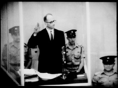 Adolf Eichmann raising a hand during a trial, standing behind glass walls and surrounded by guards.