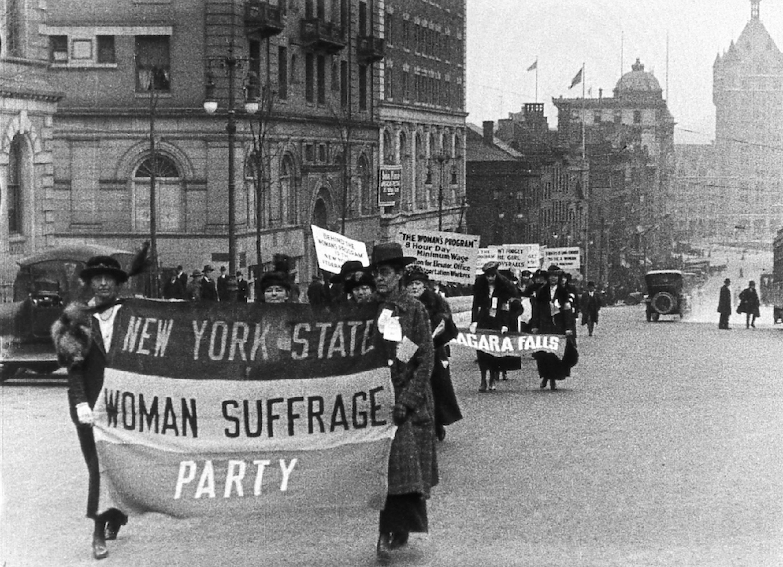 Women marching in the street with a banner for the New York State Woman Suffrage Party.