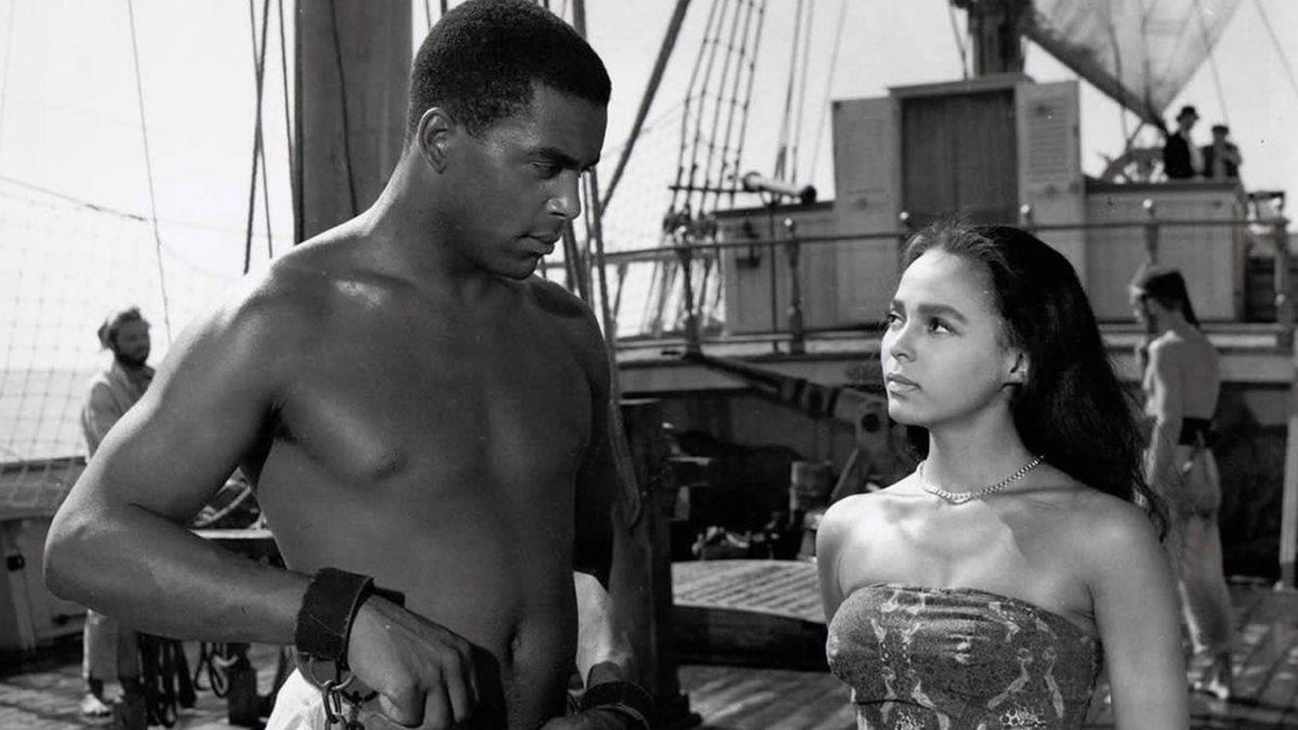 A man and woman looking at each other on a ship. The man is enslaved in shackles.
