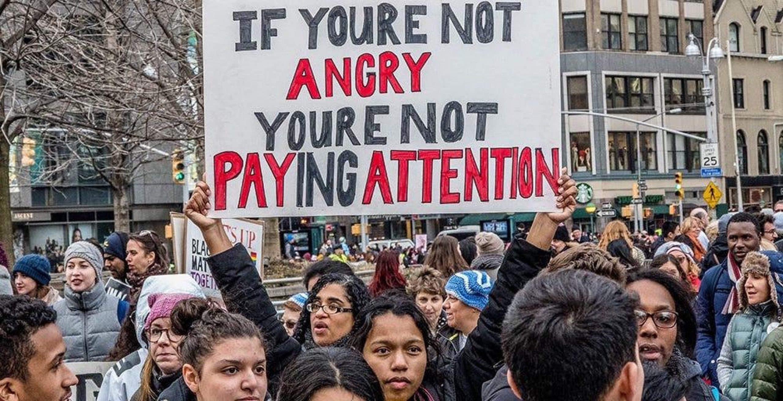 crowd of people with one woman holding up a sign "if you're not angry you're not paying attention"