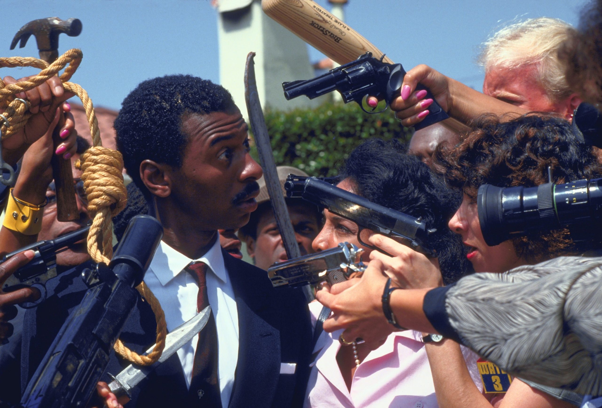 Still from "Hollywood Shuffle." Robert Townsend as character Bobby Taylor. Press and paparazzi crowd with microphone, hammer, camera, gun to face.