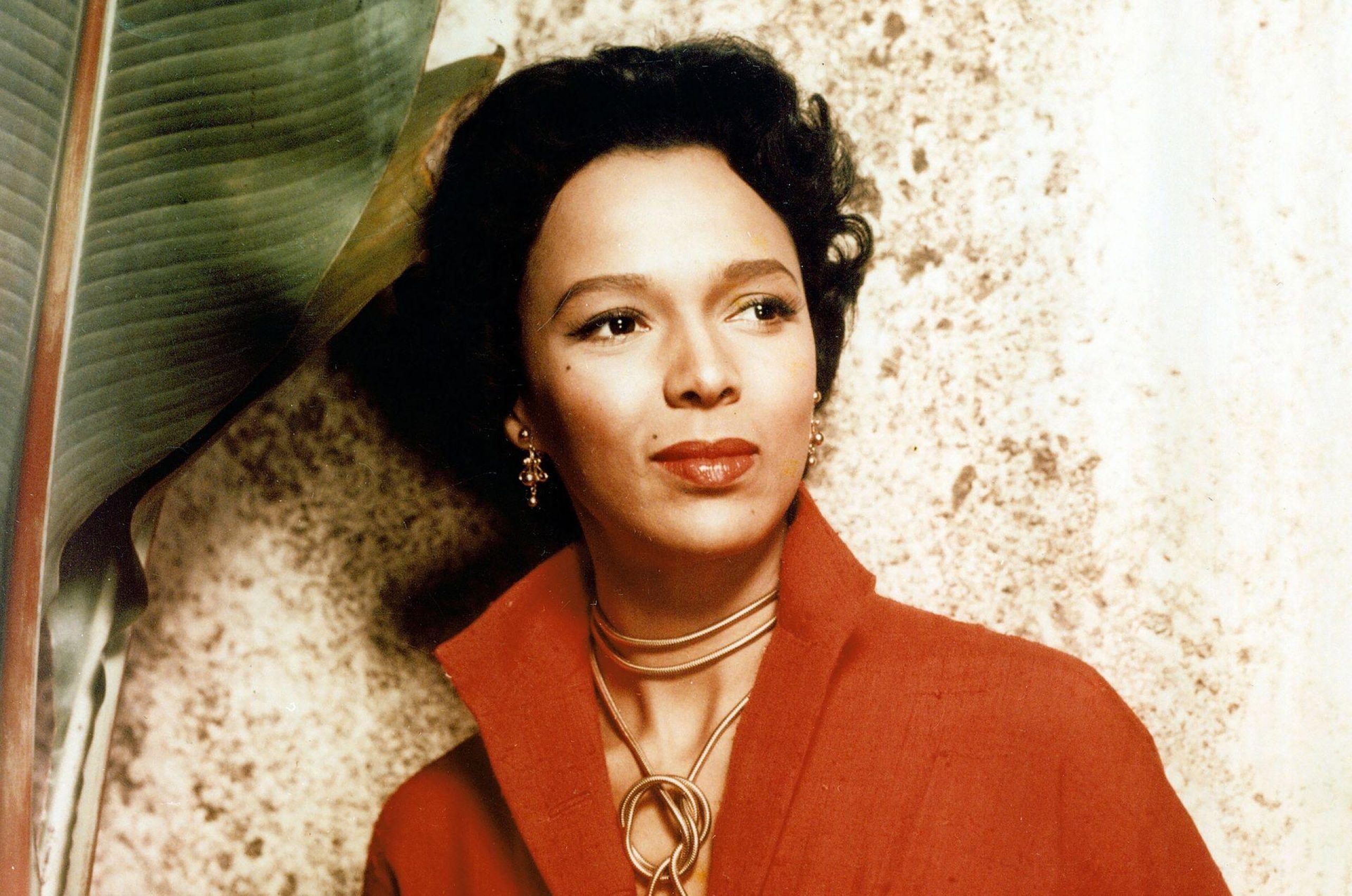Dorothy Dandridge in a red outfit and gold jewelry poses beside a large tropical leaf against a textured wall.