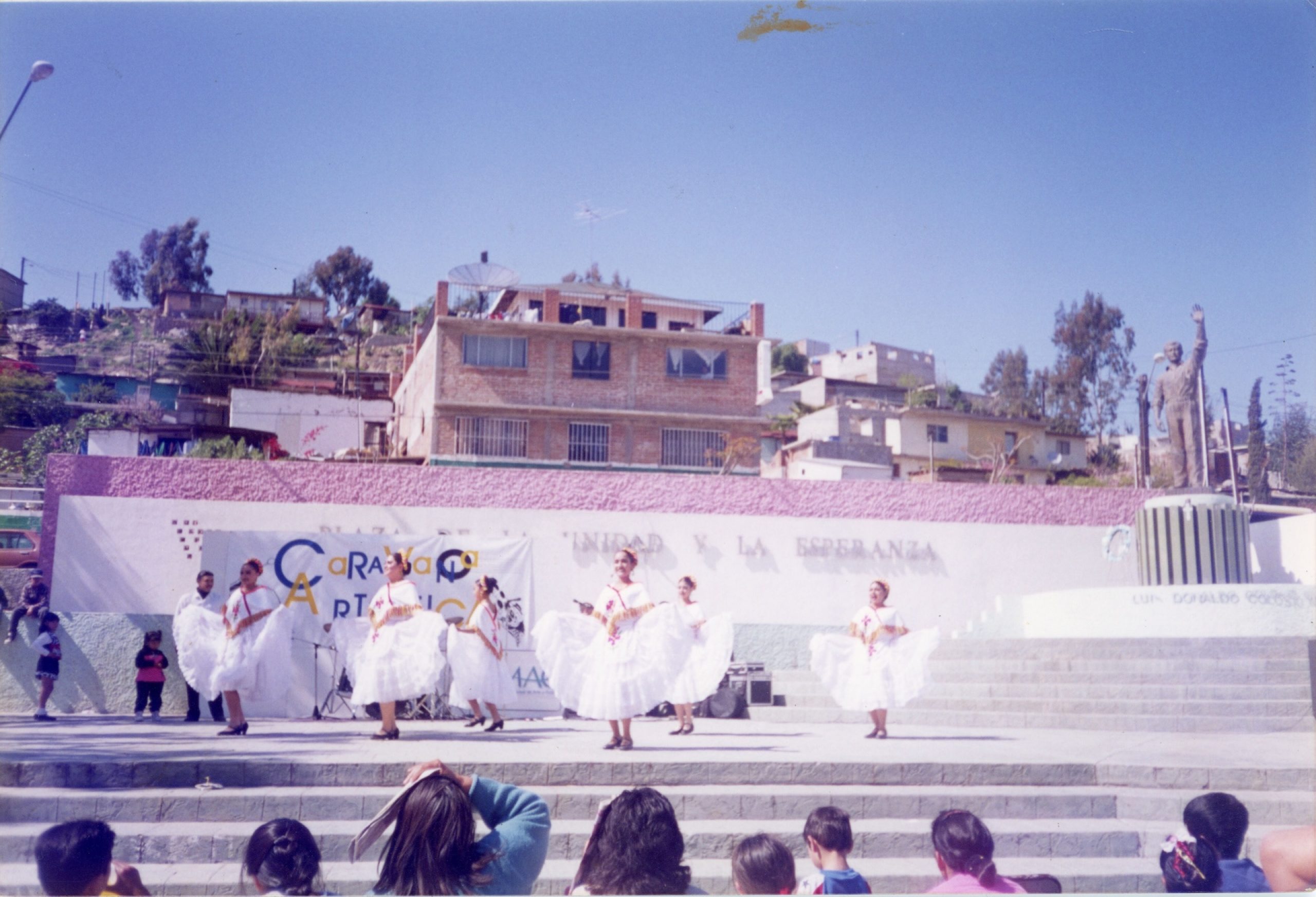 Dancers in white folklórico dresses performing at an outdoor plaza event, with hillside homes in the background.