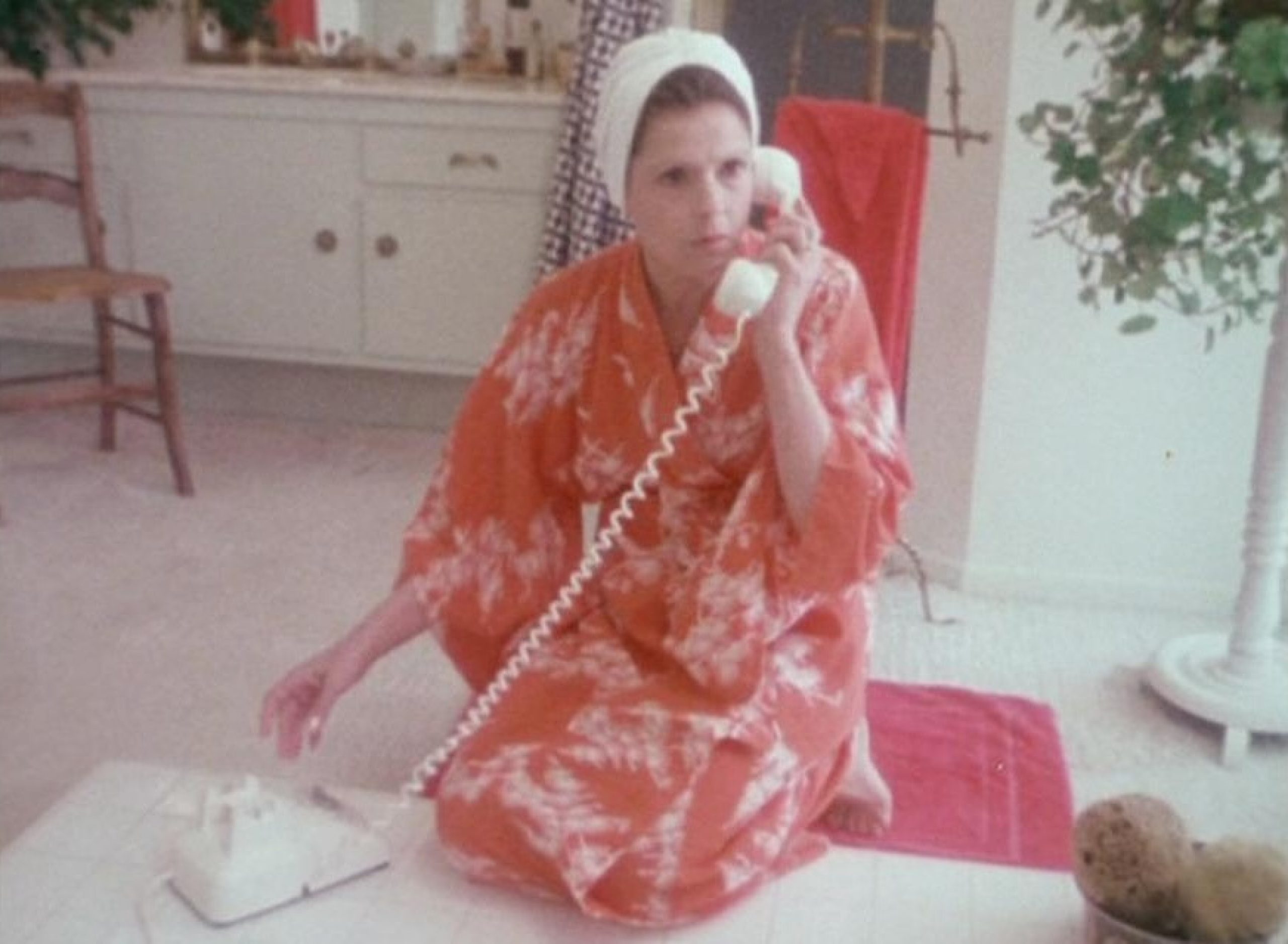 Woman in pinkish orange robe and head covered holding a phone receiver to her ear