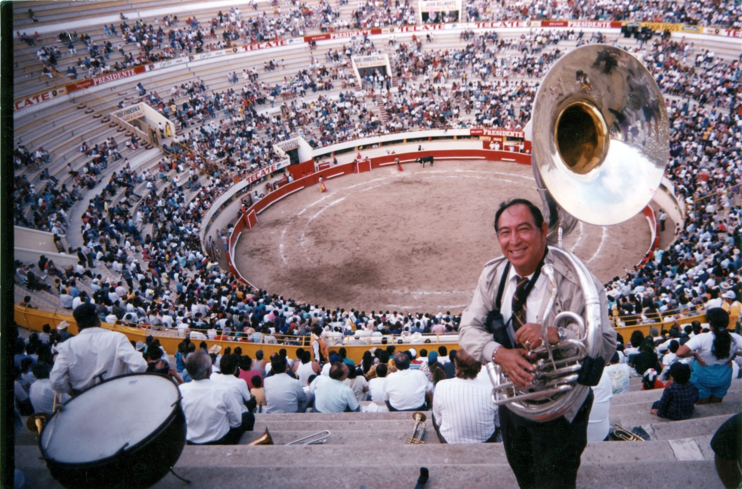 Man holding a sousaphone smiling for the camera, with a packed bullring and matador visible below.