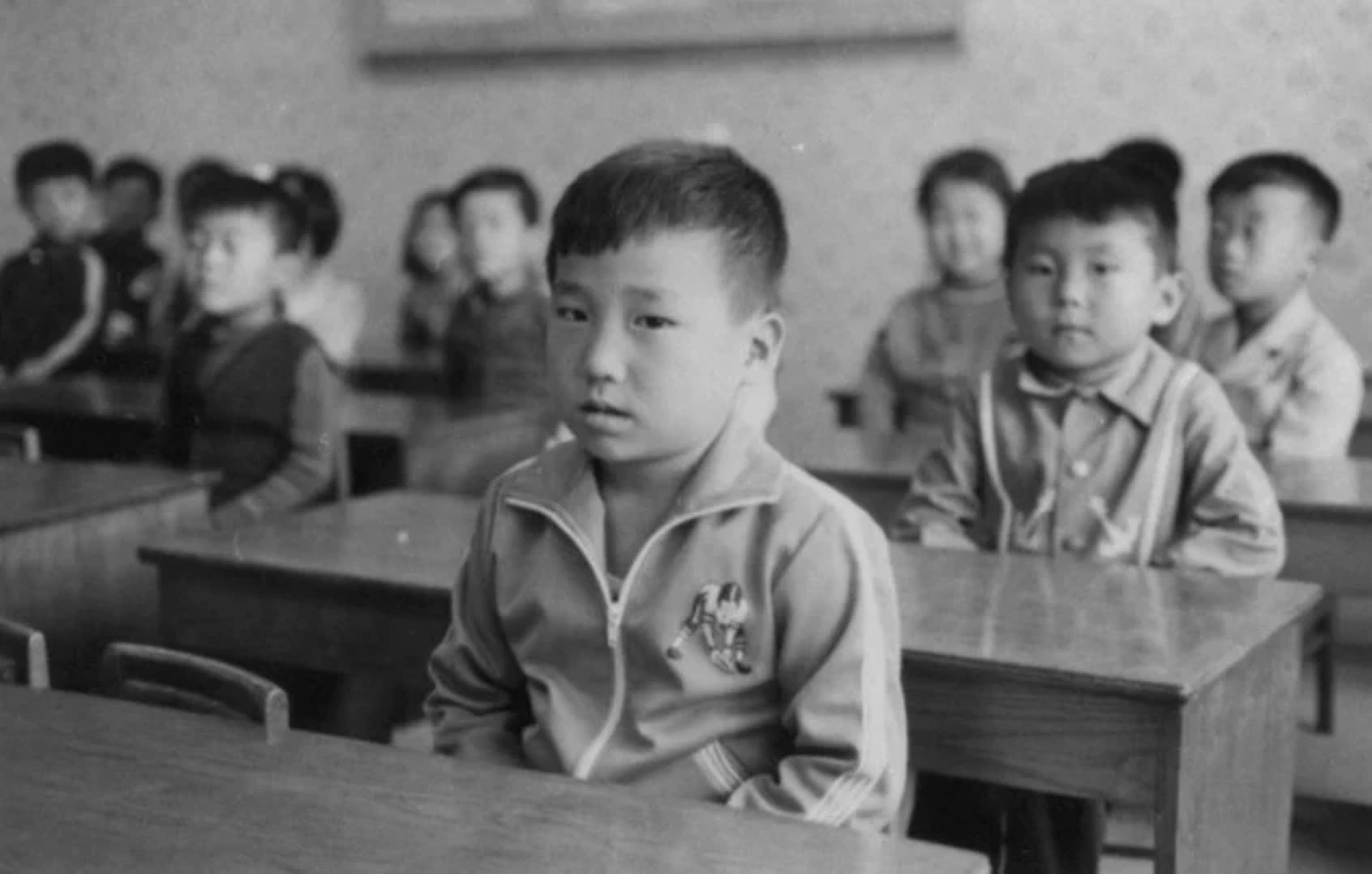 young boy sitting at a desk with other children at desks in the background
