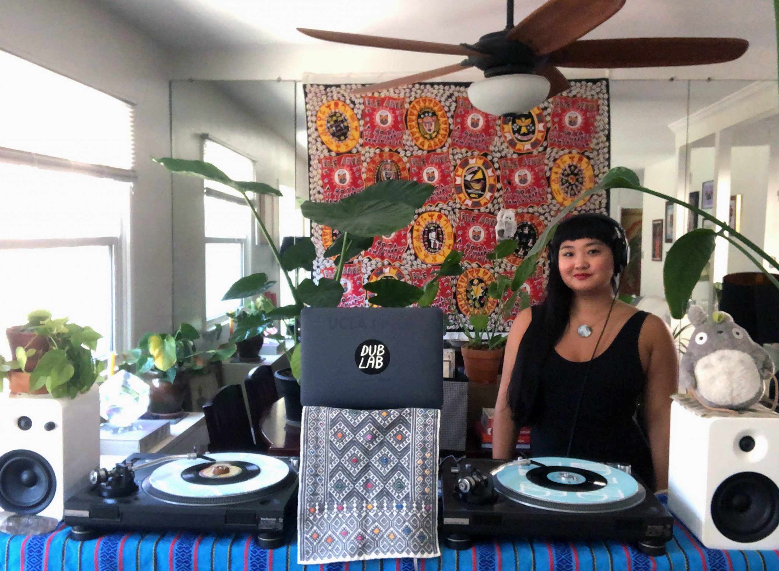 A woman with black hair and black tank top standing behind turntables, smiling at the camera