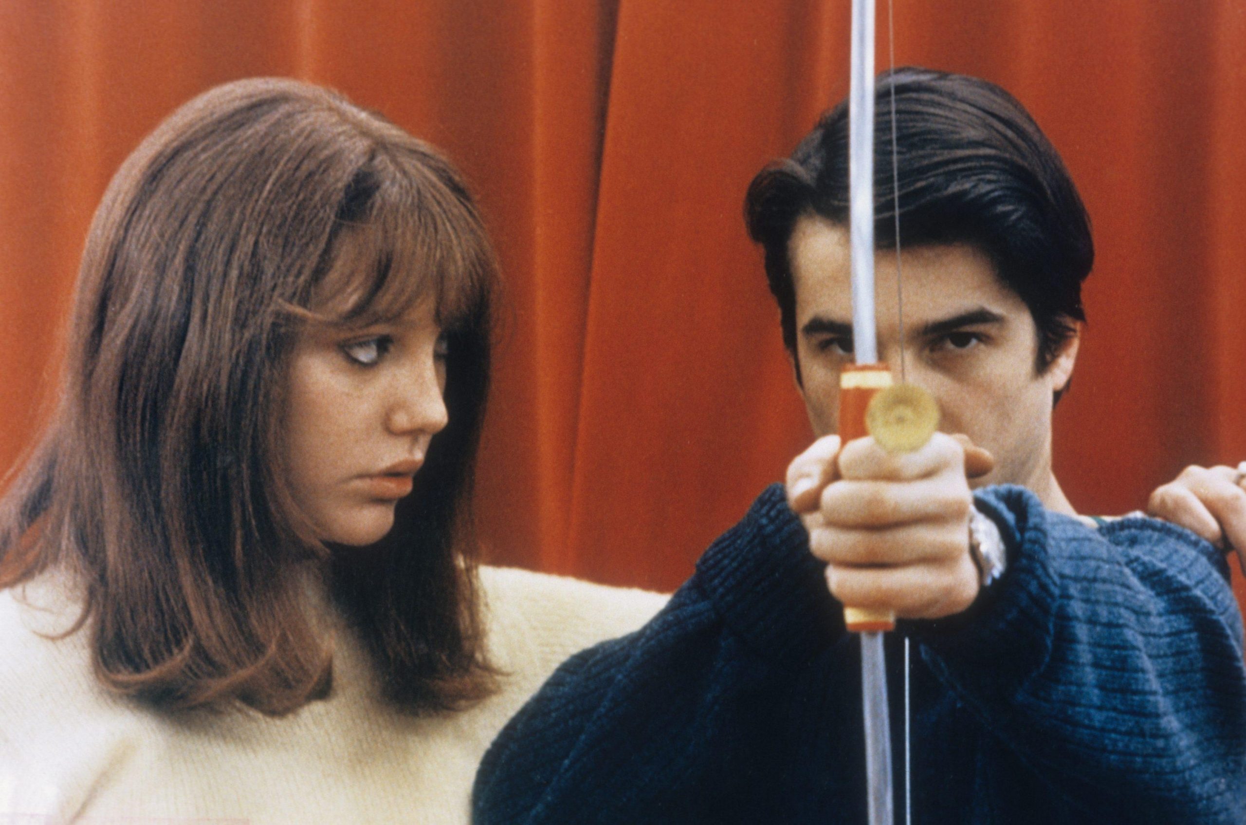 A woman watching a man aim a crossbow at the camera