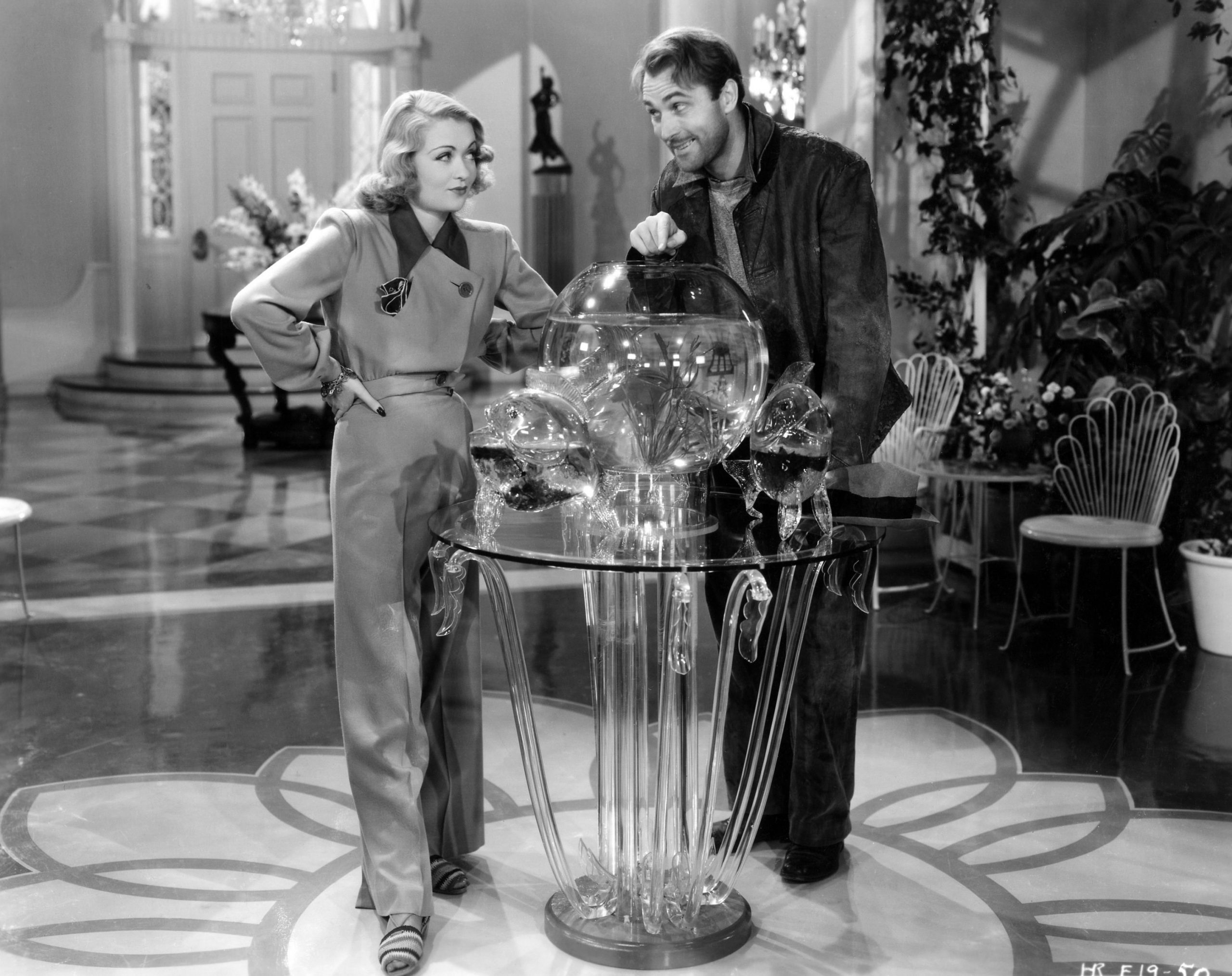 woman in pantsuit and man in disheveled clothes standing behind a glass table