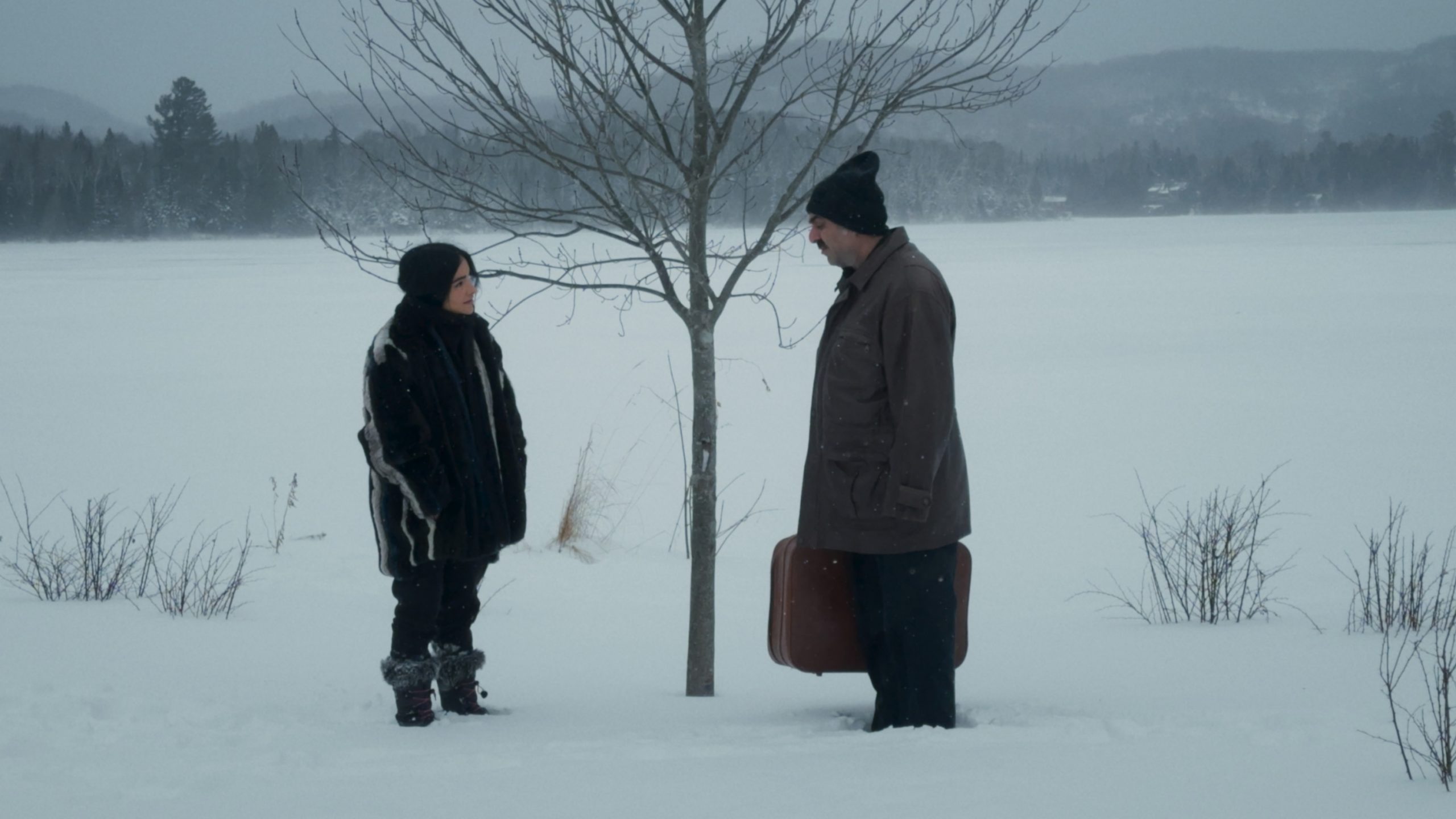 man dressed warmly and holding a suitcase talking to another person with both standing in the snow