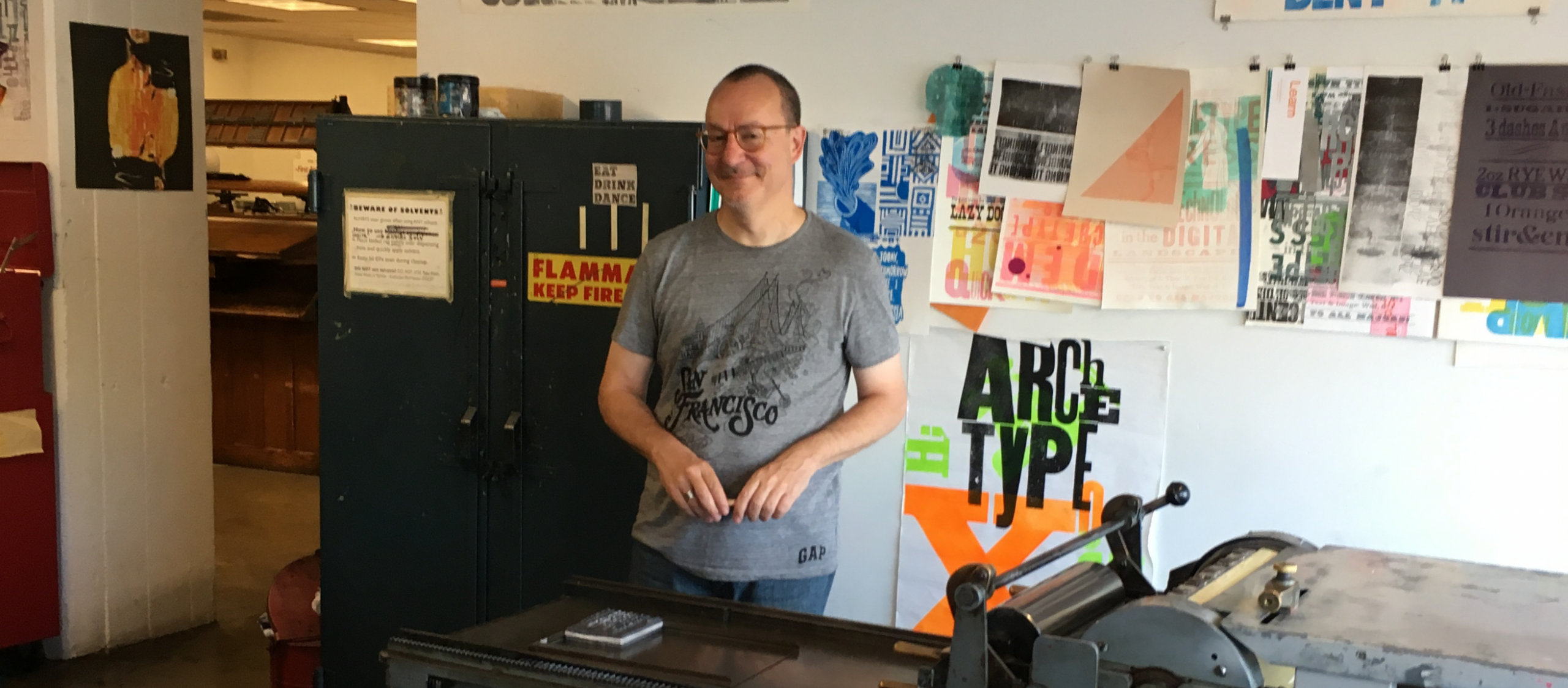 Robert gore in t-shirt standing in front of posters and behind a printing press