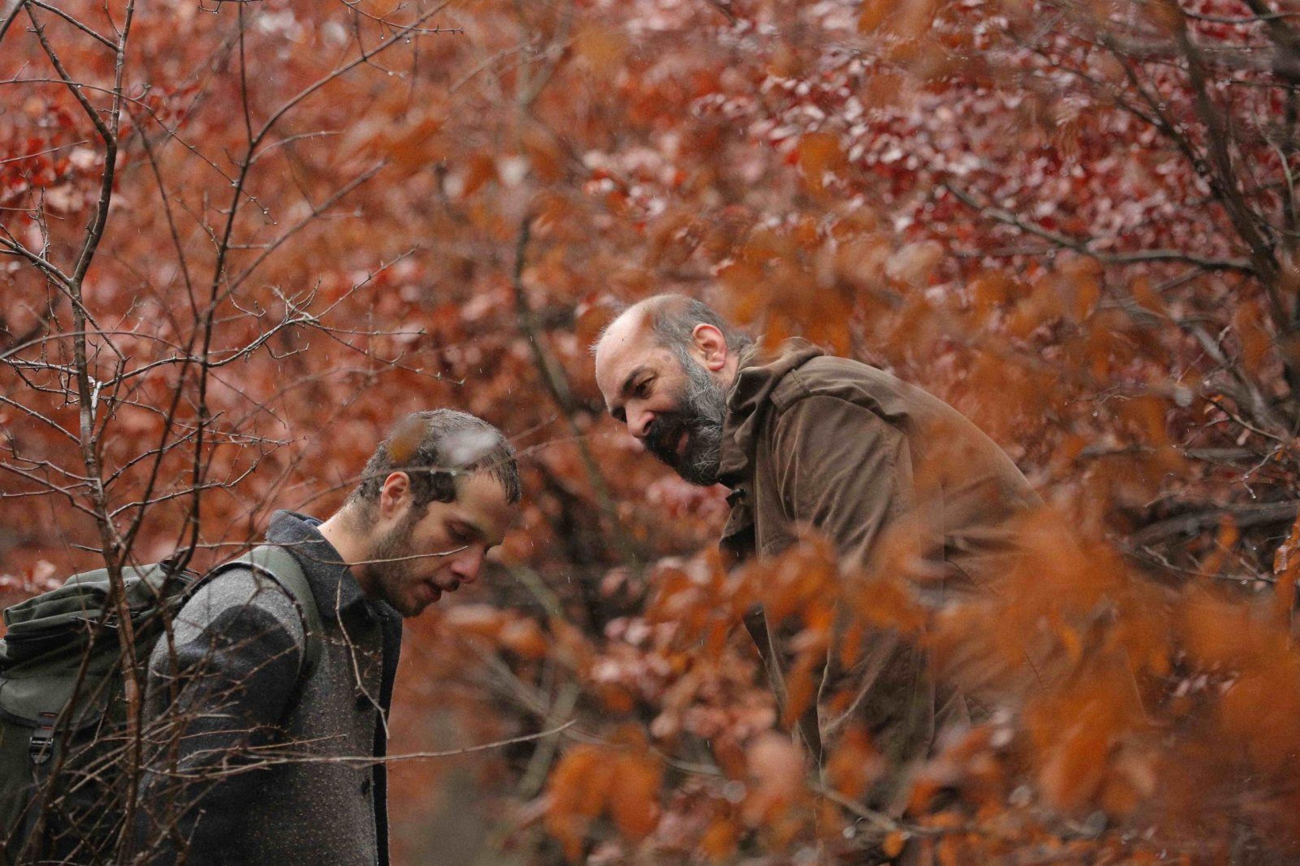 Two men face each other heads down in a wooded area surrounded by thin tree branches with red leaves