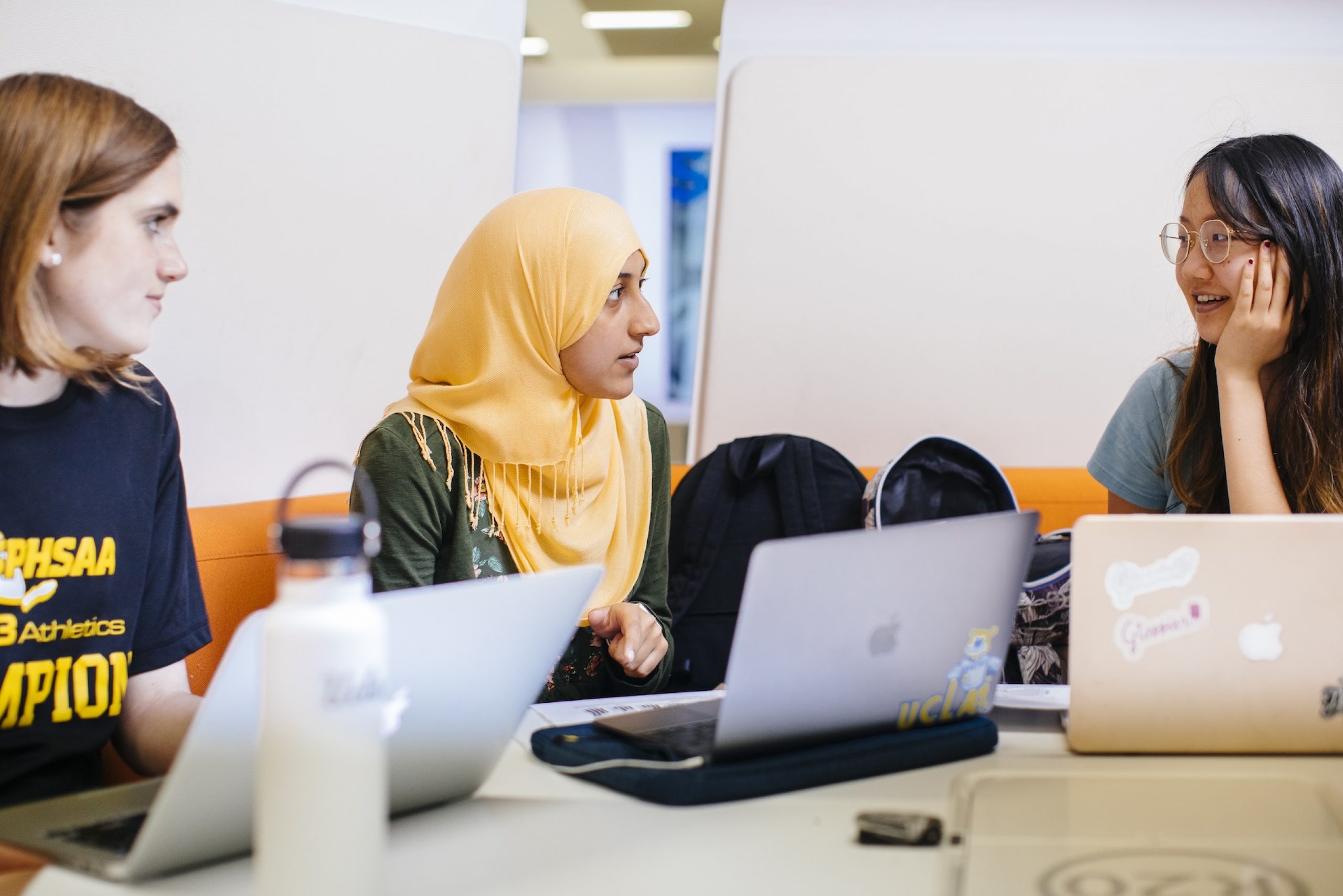 Three students sitting at a table while on their computers and talking