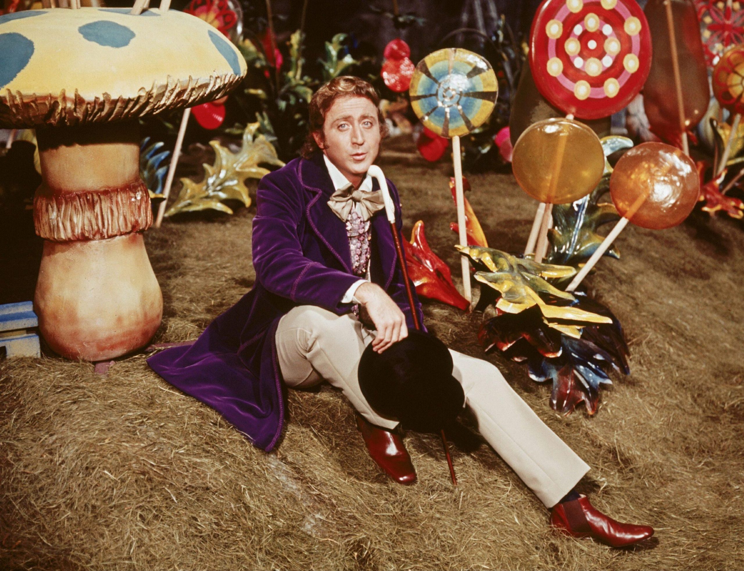 A man (willy wonka) in a purple suit sitting amongst large candies