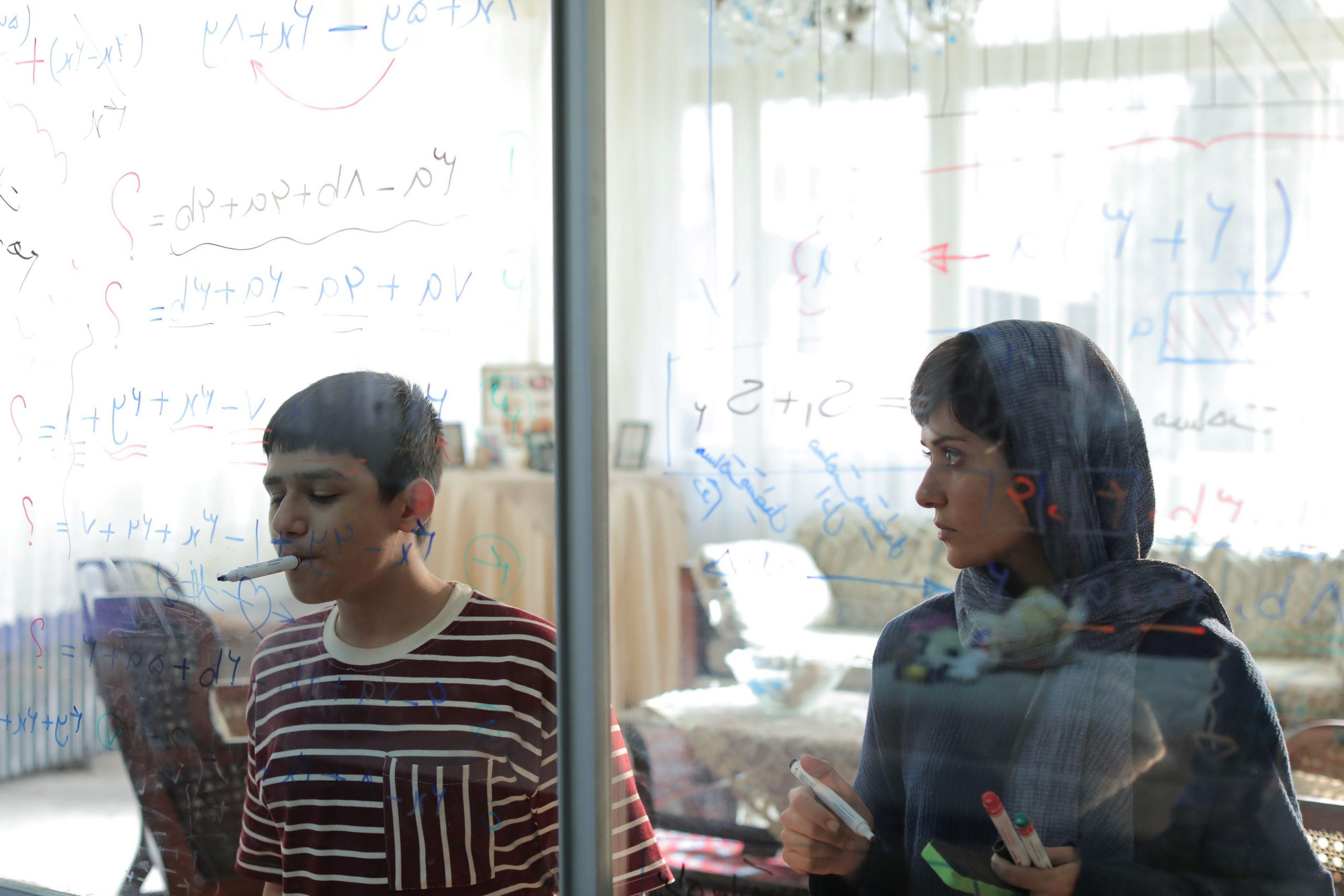 boy with marker in his mouth writing on clear glass while woman in a headscarf stands next to him and looks on