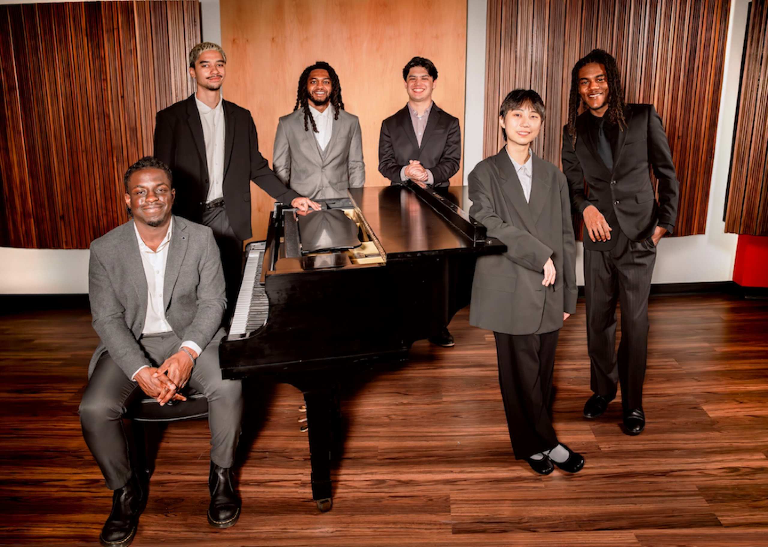six members of the Herbie Hancock student ensemble wearing grey suits and standing around a grand piano