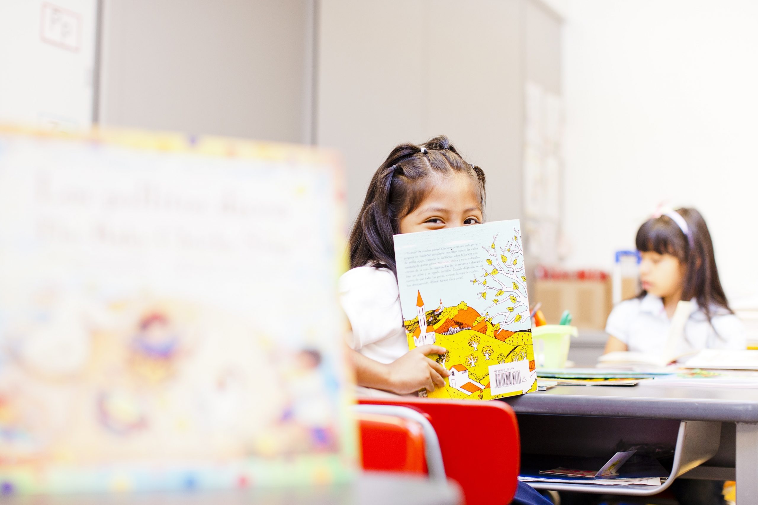 young girl sitting at a desk peeking over a book