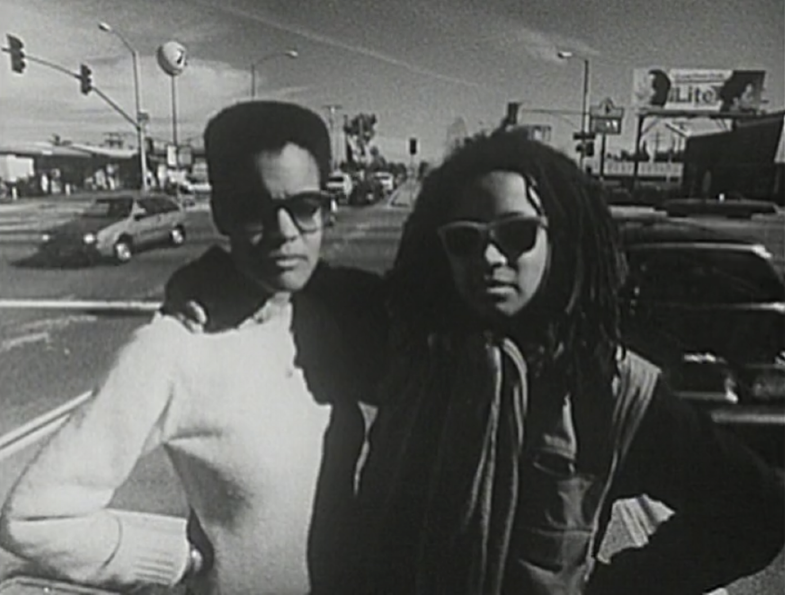 Person in white with hair cropped close in white top and person with dreads and arm around person in white staring to the front
