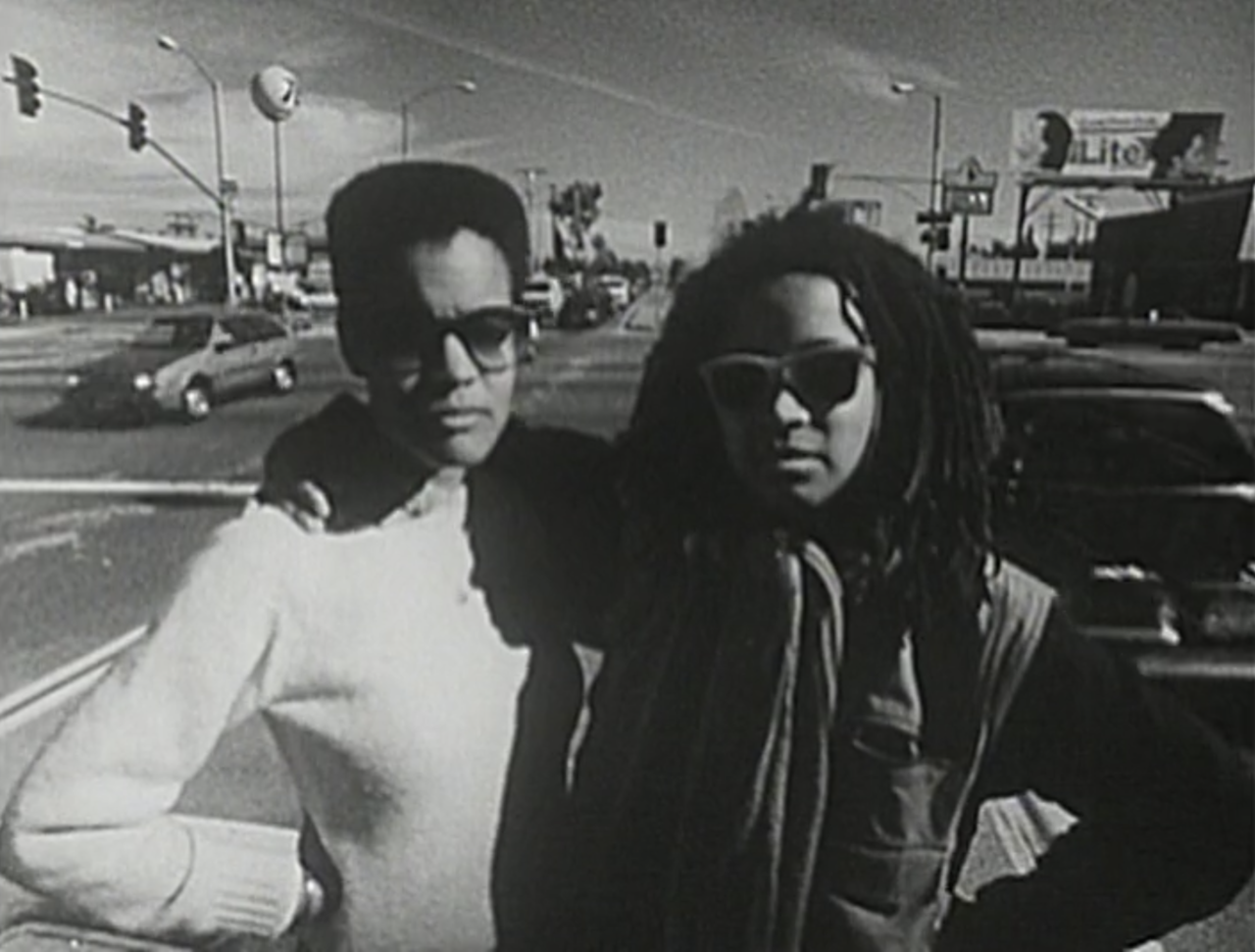 Person in white with hair cropped close in white top and person with dreads and arm around person in white staring to the front
